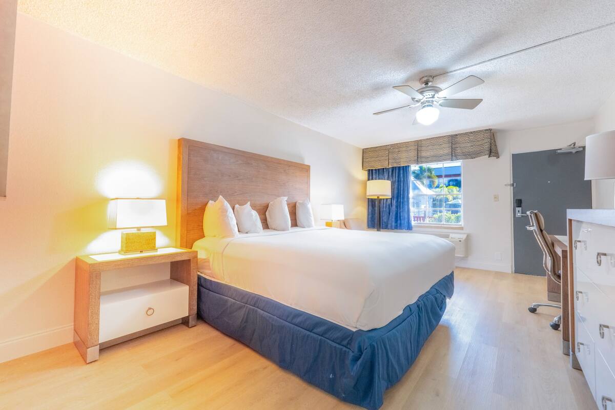 Hotelkamer met een groot bed, nachtkastje met lamp, plafondventilator, raam met gordijnen en een dressoir.