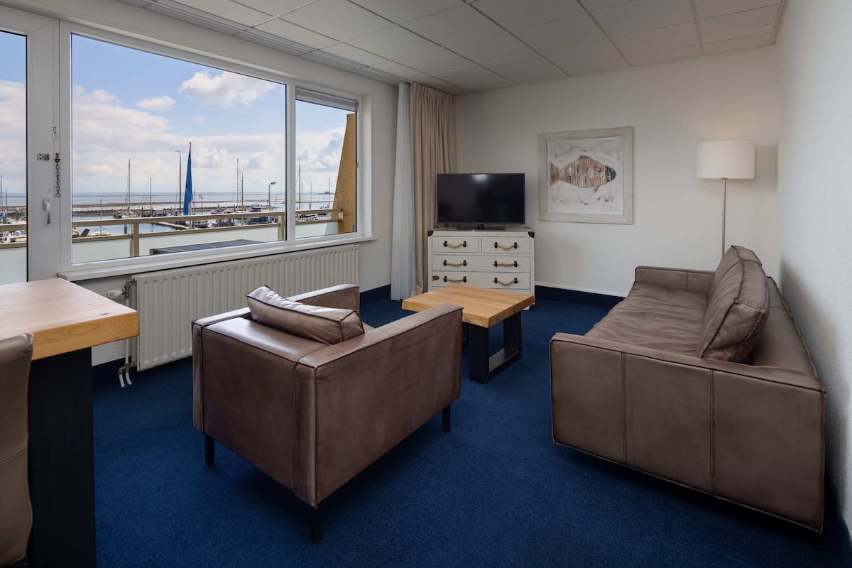 Hotelkamer met een groot raam dat uitzicht biedt op een jachthaven, een bank, een salontafel, een televisie en een lamp.