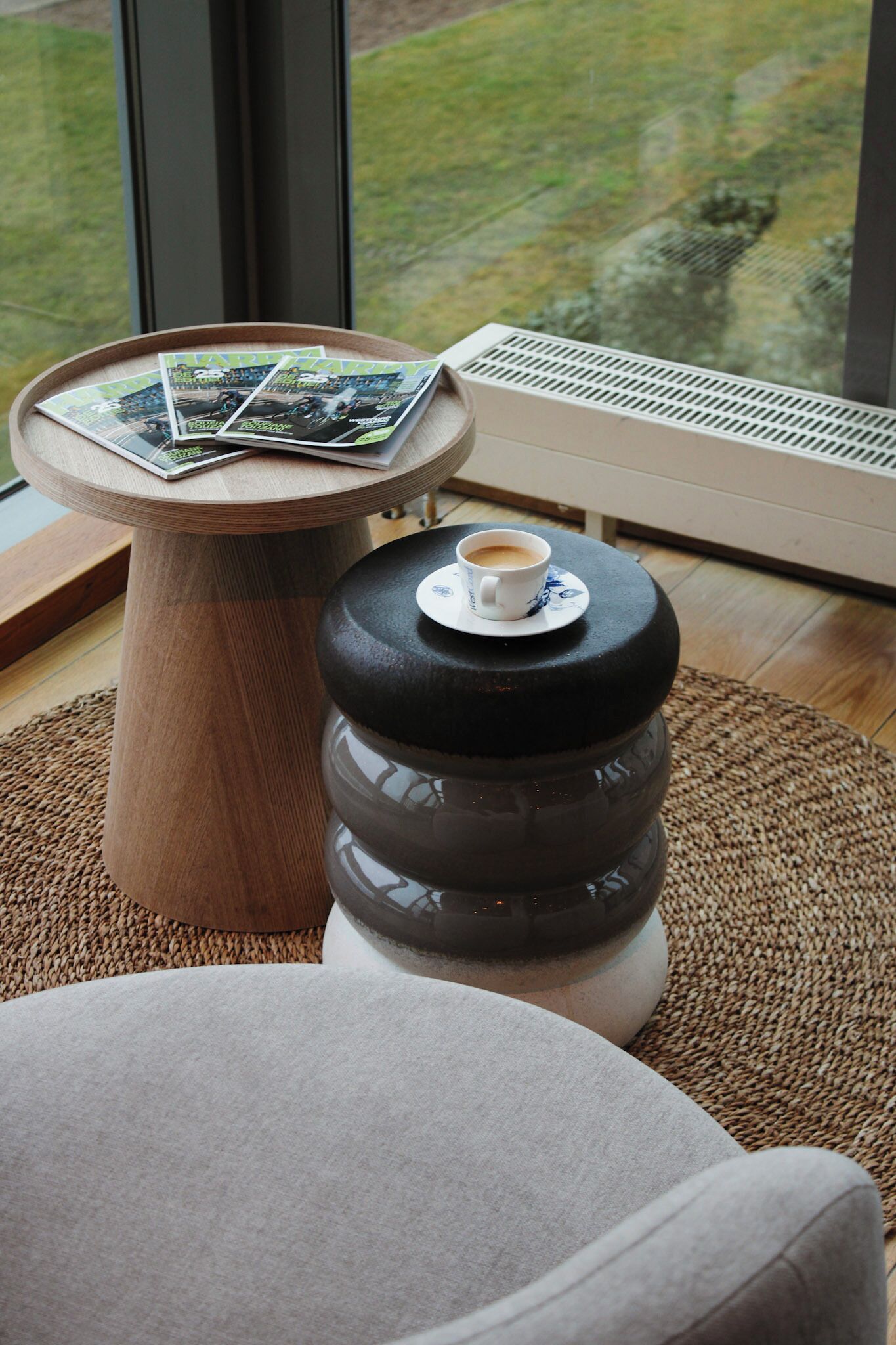 A cozy room with a round wooden table holding magazines and a cup of coffee, a black ottoman with a coffee cup, and a large window with a view of a grassy area.