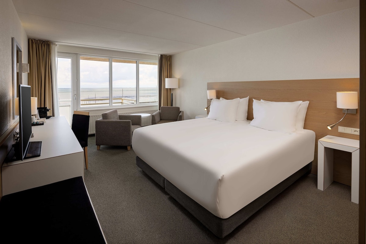 Een hotelkamer met een groot bed, een bureau met een computer, een stoel, een klein tafeltje en een raam met uitzicht op zee.