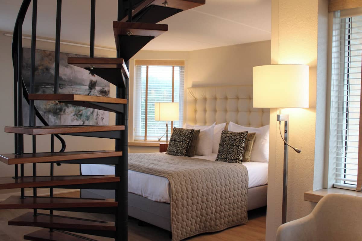 Slaapkamer met een bed, een bedlamp, een stoel, een raam met zonwering, en een trap met een schilderij.