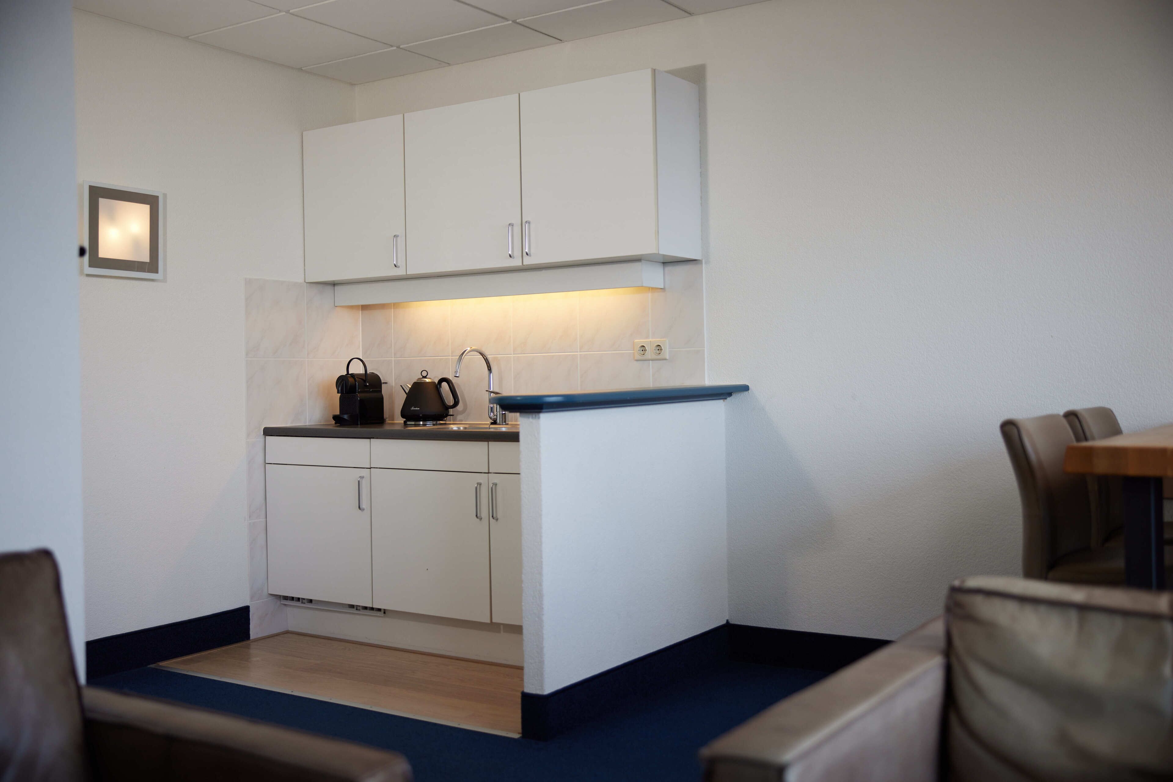 A compact kitchen with white cabinets, a countertop, and a kettle. There are chairs and a table in the adjacent dining area.