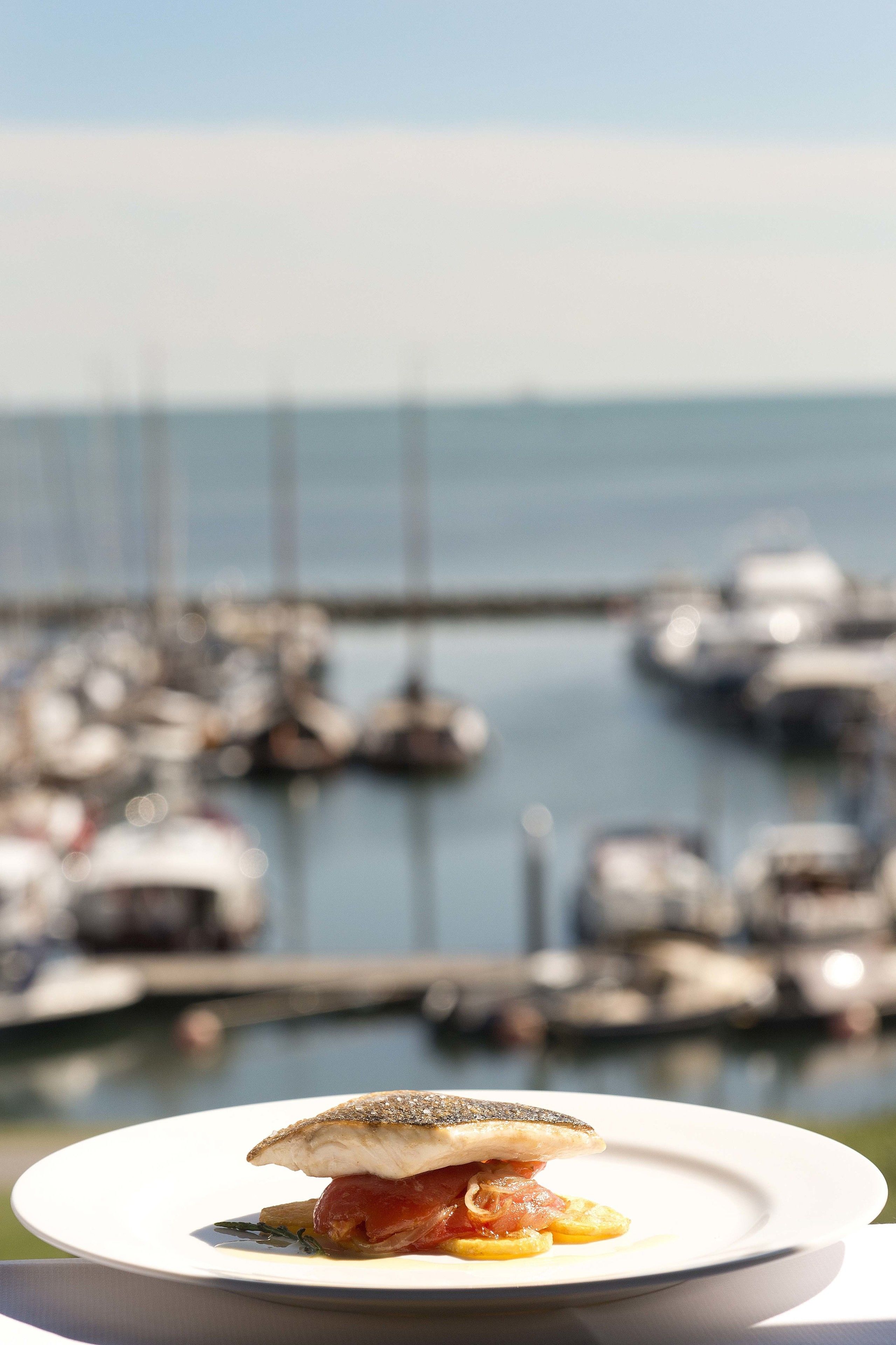 A plate of food with a fish and vegetables, set against a marina with boats in the background.