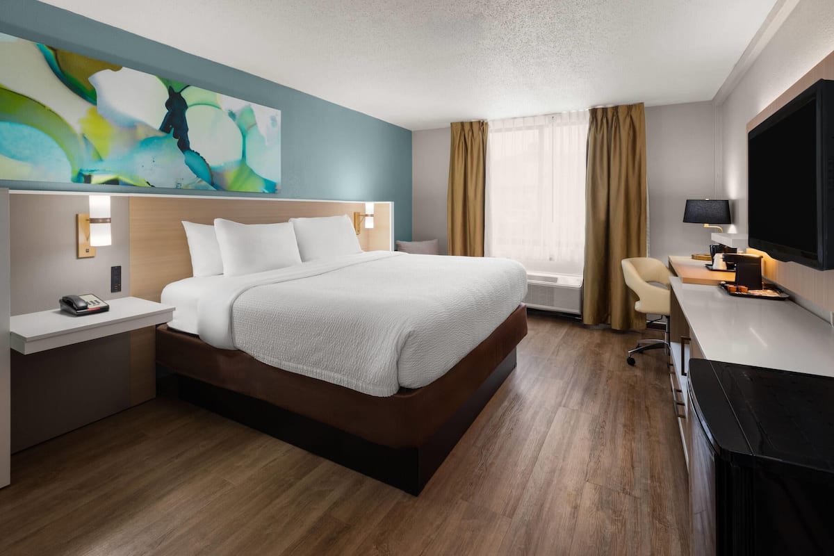 A hotel room with a large bed, a desk with a flat-screen TV, a wall-mounted TV, and a painting on the wall.
