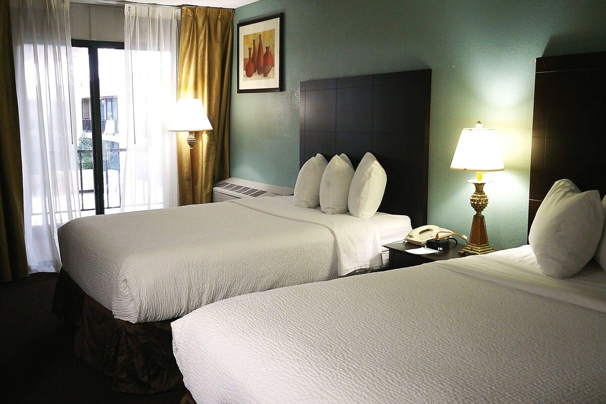 A hotel room with two beds, a nightstand with a lamp, a phone, and a window with curtains.