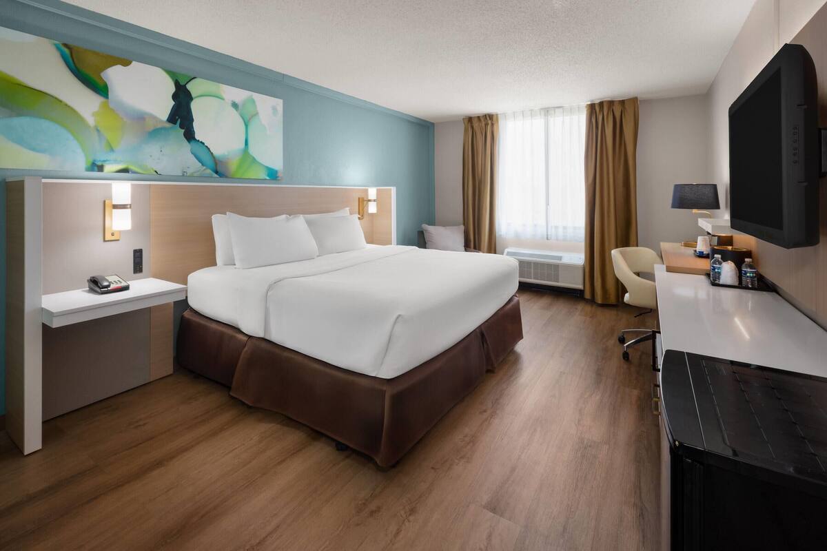 A hotel room with a large bed, bedside tables, a desk, a chair, a flat-screen TV, and a painting on the wall.