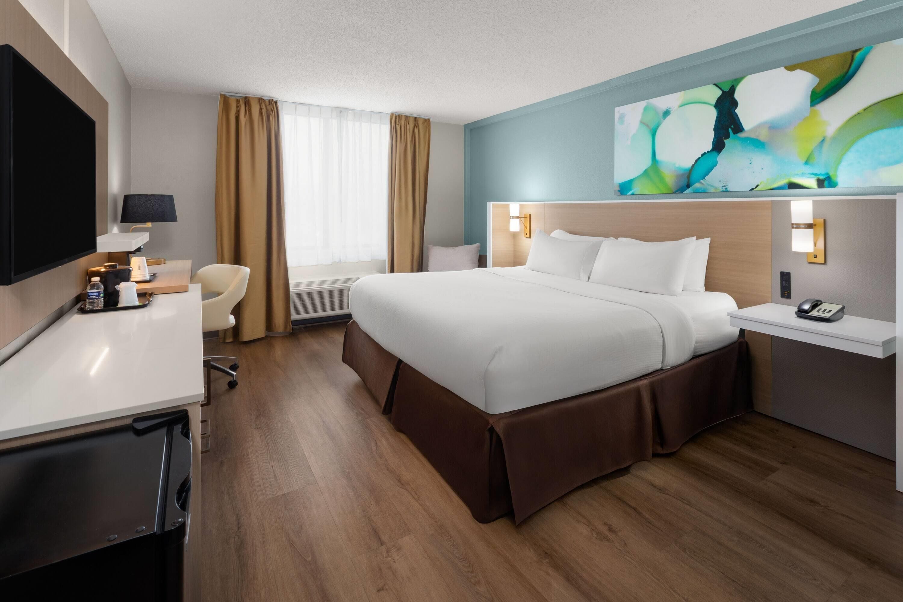 A hotel room with a bed, a desk, a chair, a TV, and a painting on the wall.
