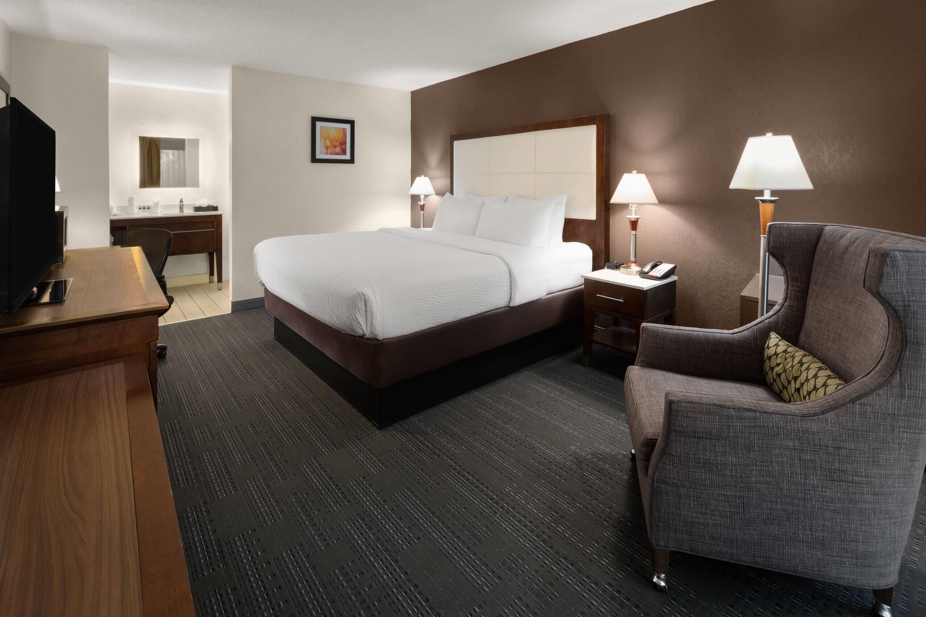 A hotel room with a bed, a desk, a chair, a TV, and a lamp.