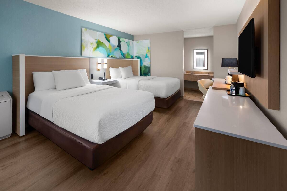 A hotel room with two beds, a desk, a chair, a TV, and abstract art on the wall.