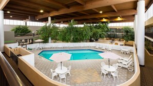 A pool area with white chairs and umbrellas, surrounded by indoor plants and a high ceiling.