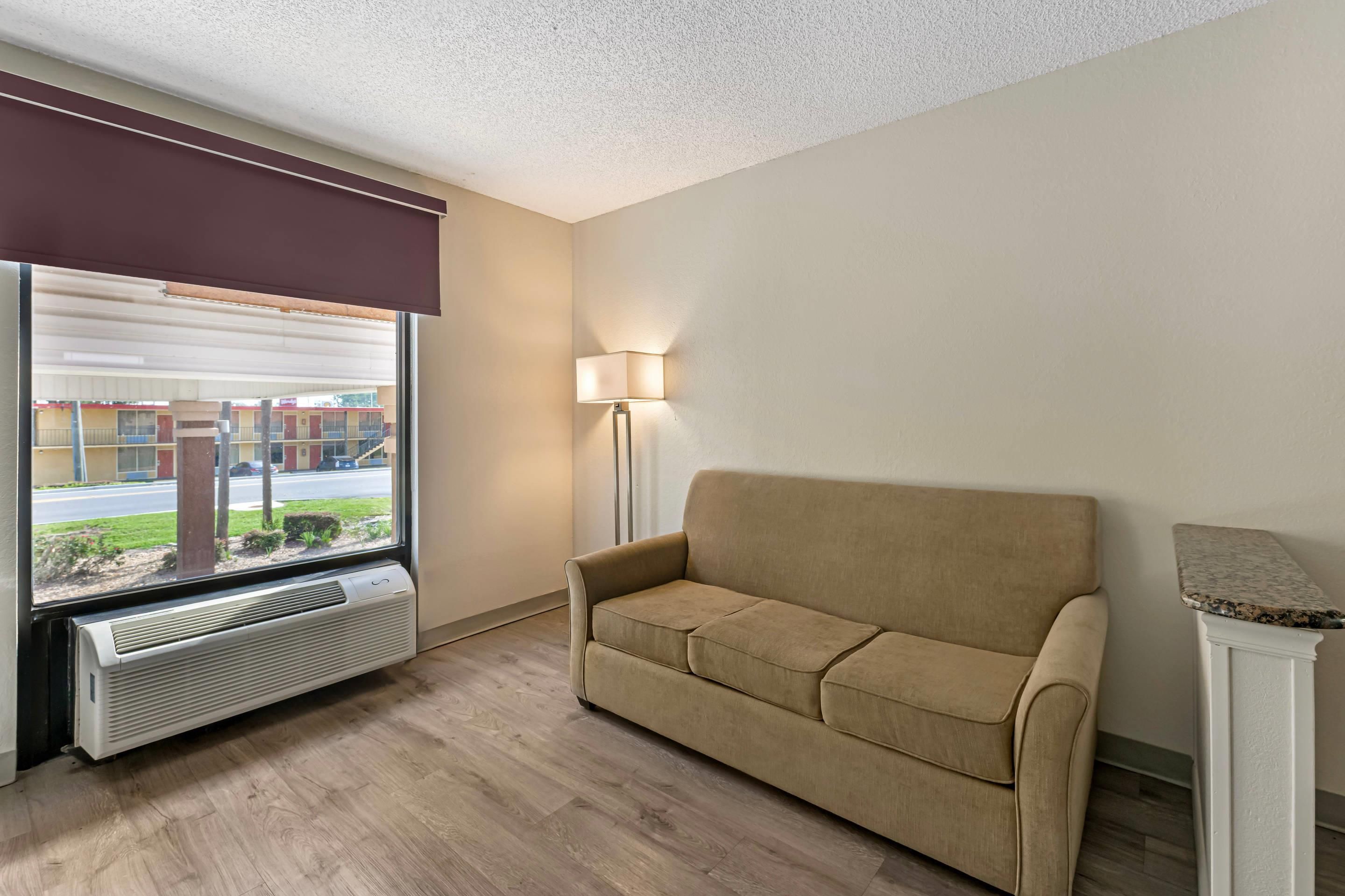A hotel room with a beige sofa, a standing lamp, a window with a view of buildings, and a wall-mounted air conditioner.