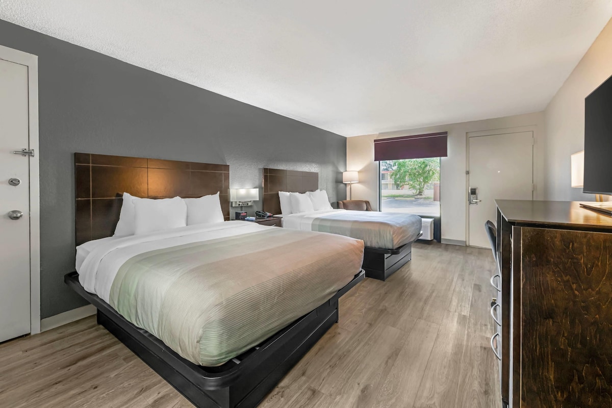 A hotel room with two beds, a desk, a chair, a lamp, and a view of the outdoors.