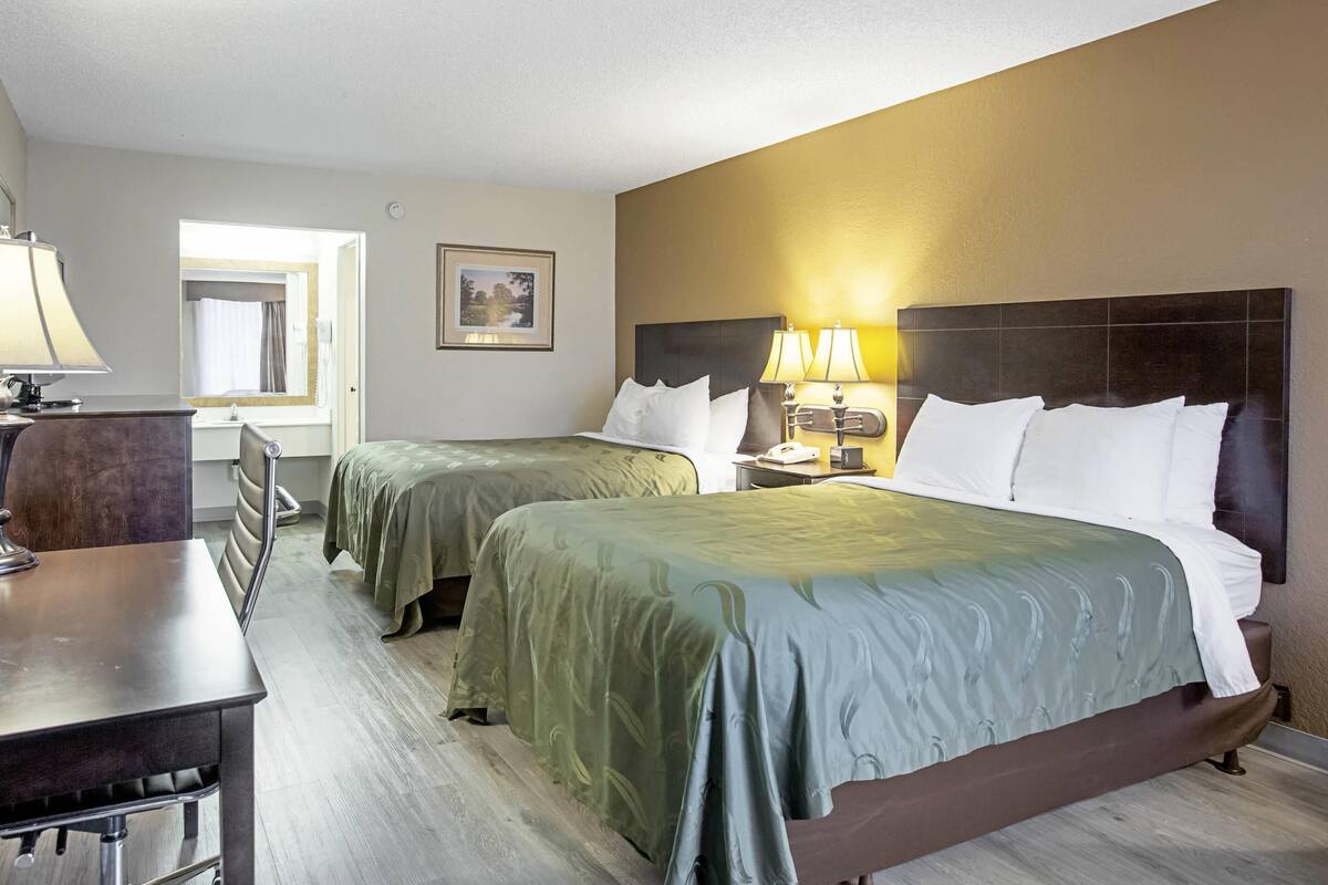 A hotel room with two beds, a desk, a chair, a lamp, and a framed picture on the wall.
