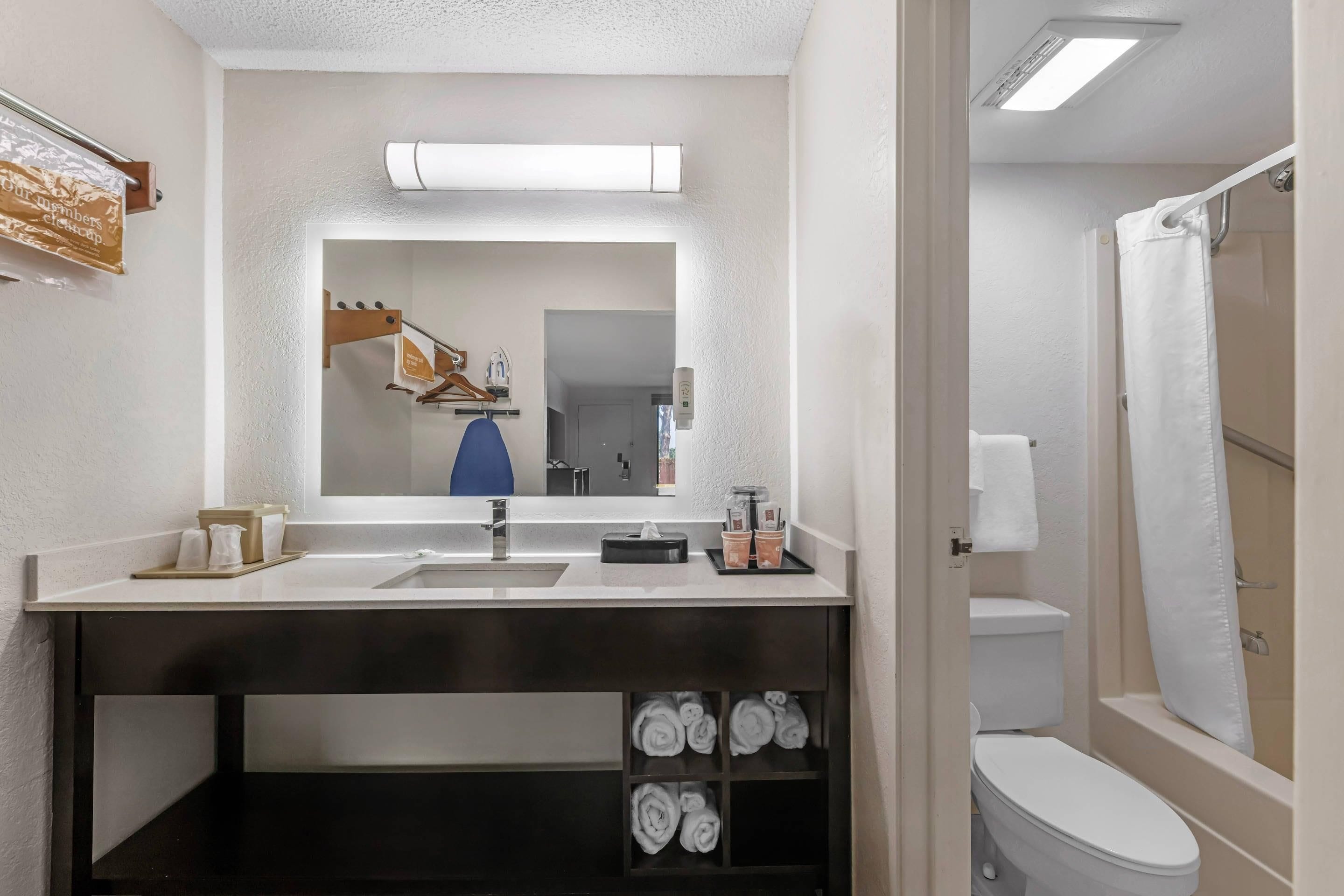 A modern bathroom with a sink, mirror, and toilet. There is a towel rack with towels and a shower curtain visible.