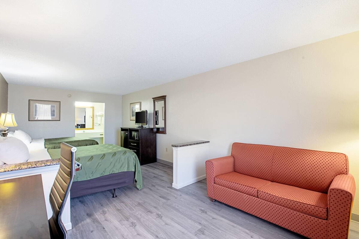 A room with a bed, a sofa, a desk with a lamp, a dresser, and a mirror.
