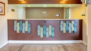 Un comptoir d’accueil avec un panneau marron et un tissu à motifs suspendu, une enseigne au-dessus indiquant « Quality Inn & Suites by Choice Hotels », et un miroir au mur.