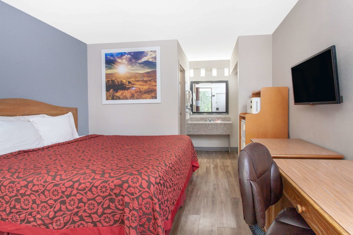 A hotel room with a bed, a desk, a chair, a TV, a window, and a bathroom visible in the background.