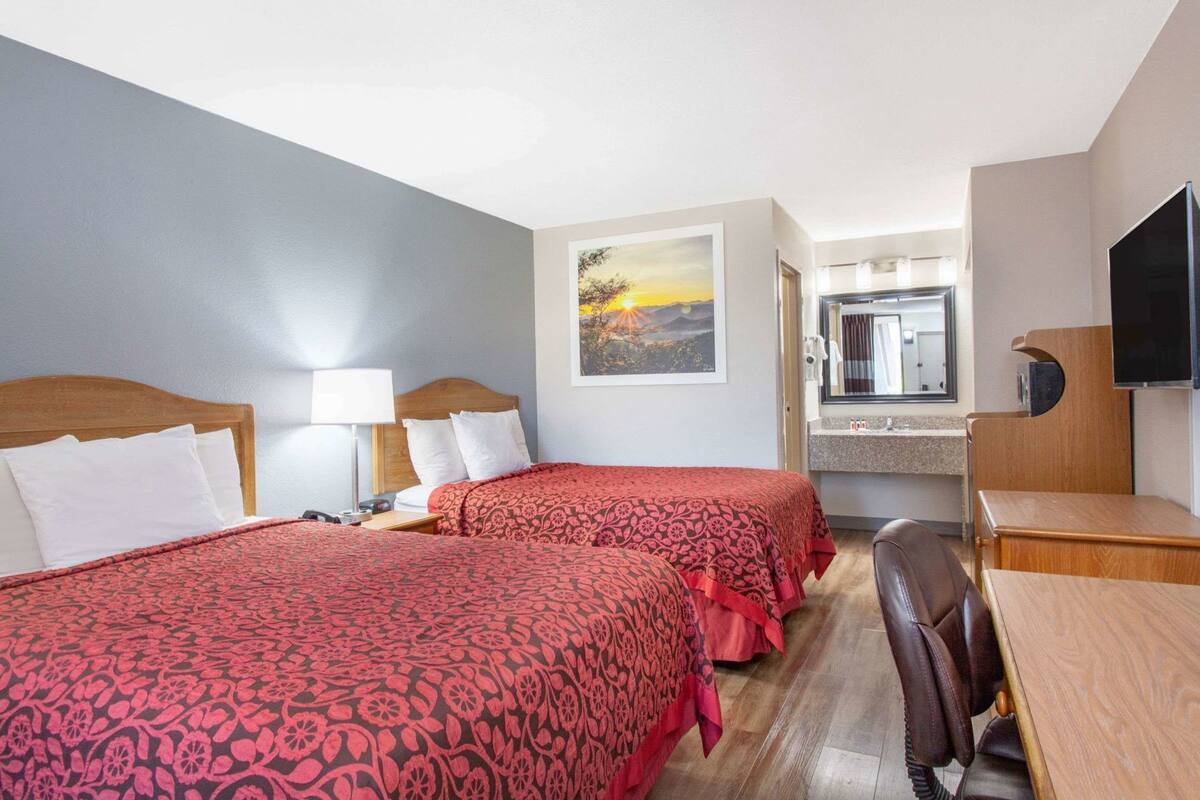 A hotel room with two beds, a desk, a chair, a TV, a bathroom mirror, and a painting on the wall.