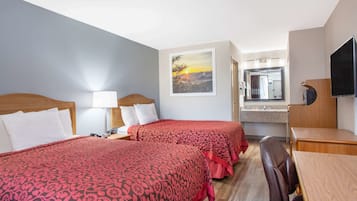 A hotel room with two beds, a desk, a chair, a TV, a bathroom mirror, and a painting of a sunset.