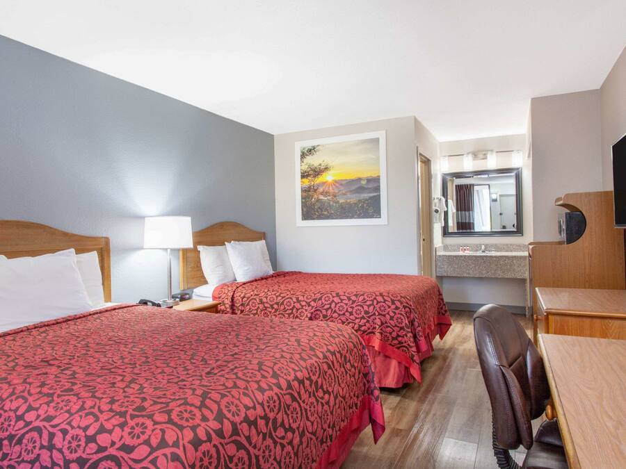 A hotel room with two beds, a desk, a chair, a TV, a bathroom mirror, and a painting of a sunset.