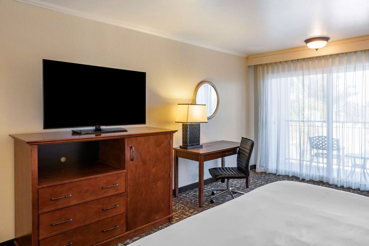 A hotel room with a large bed, a TV on a wooden cabinet, a desk with a chair, a lamp, and a window with sheer curtains.