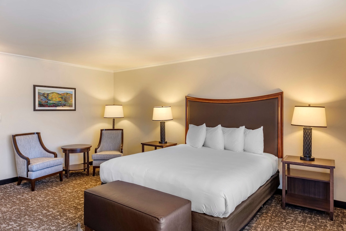 Ein Hotelzimmer mit einem großen Bett, zwei Sesseln, einem kleinen Tisch und einem Nachttisch mit Lampe.