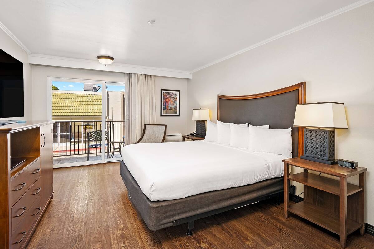 A hotel room with a large bed, a bedside table with a lamp, a TV, and a view of the outside balcony.