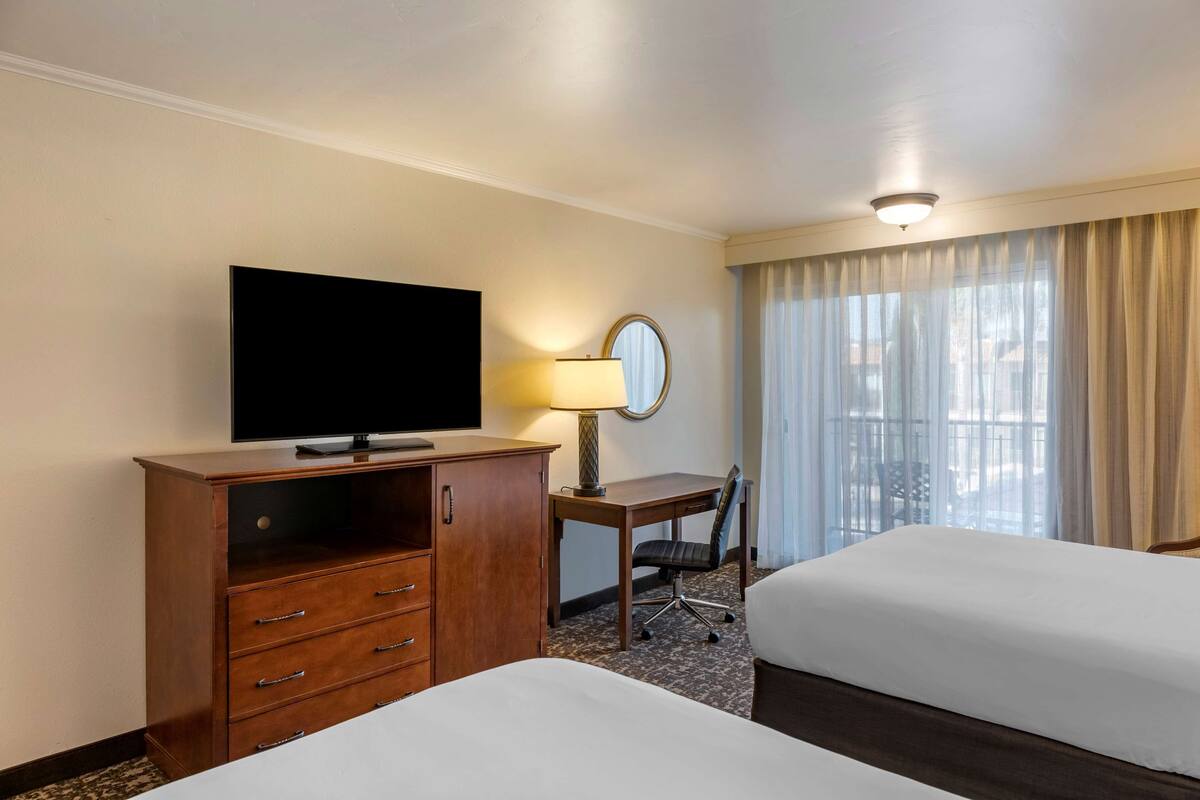 A hotel room with two beds, a TV, a desk with a lamp, and a window with curtains.