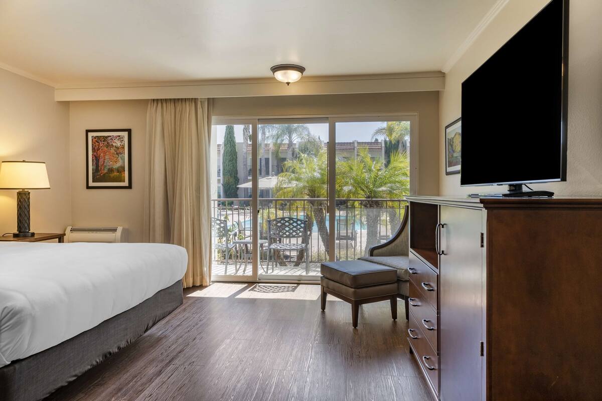 A hotel room with a bed, a TV, a desk, and a balcony with outdoor furniture.