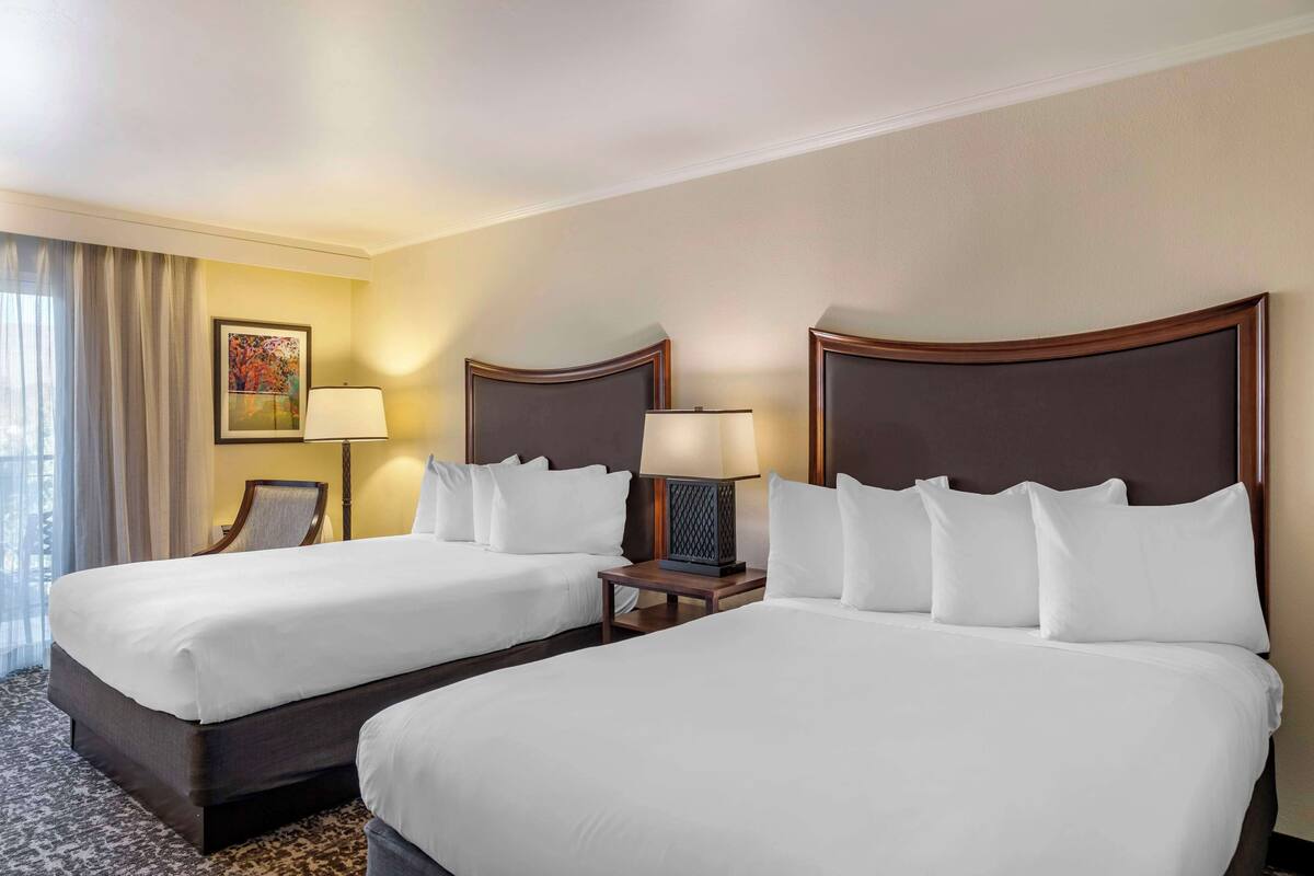 A hotel room with two beds, a nightstand with a lamp, a chair, and a painting on the wall.