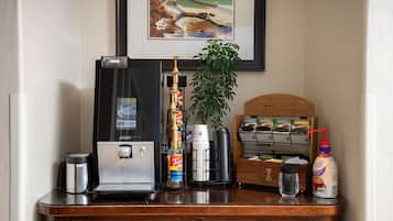 Un mostrador de recepción de hotel con una cafetera, una botella de sirope Torani, una botella de crema para café, una planta y un cuadro en la pared.