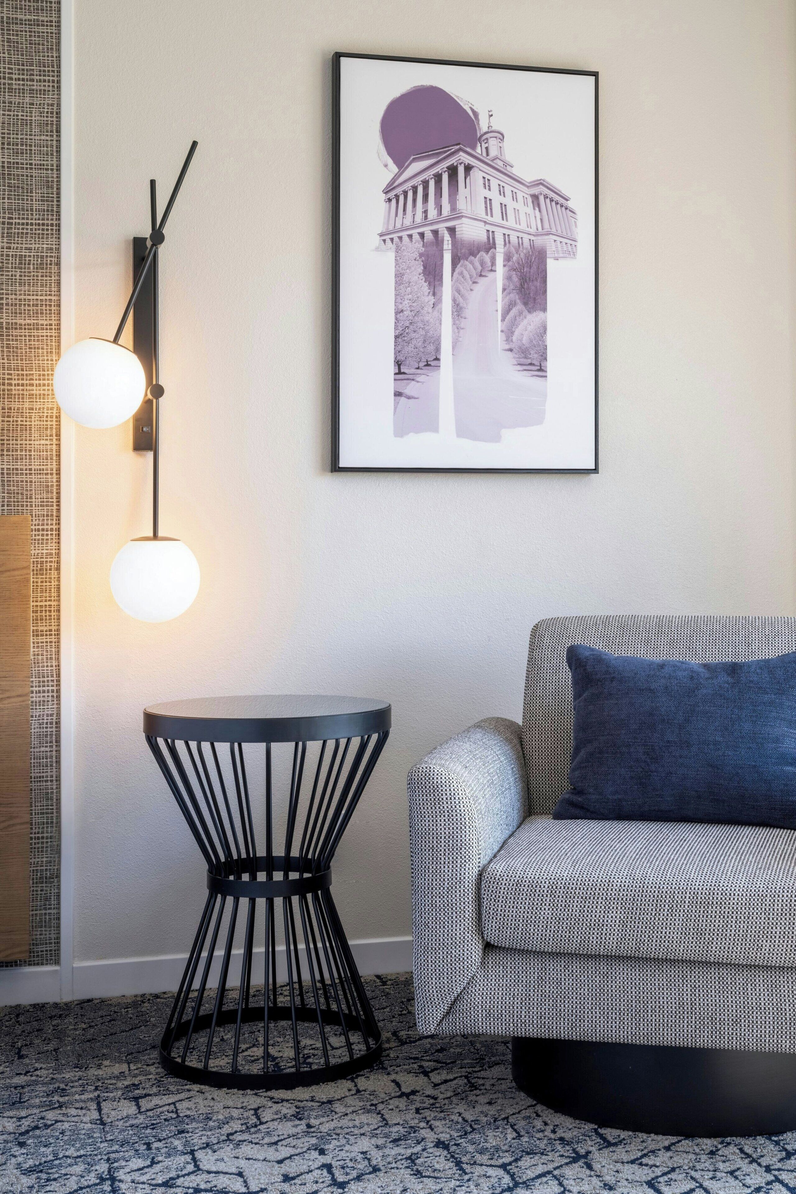 Ein modernes Wohnzimmer mit einem runden schwarzen Tisch, einem grauen Sofa mit blauem Kissen, einem gerahmten Bild eines Gebäudes und einer schwarzen Stehlampe mit zwei runden Leuchten.