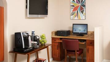 A hotel room with a desk, a computer, a coffee machine, a TV, and a colorful abstract painting.