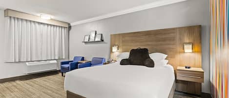 Ein Hotelzimmer mit einem großen Bett, einem Schreibtisch mit Stuhl, einem Nachttisch mit Lampe und einem an der Wand befestigten Regal mit Bildern.
