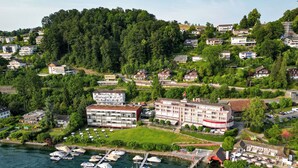 An aerial view of a coastal town with a marina, several buildings, and a forested hill in the background. - HERMITAGE Lake Lucerne - Beach Club & Lifestyle Hotel (Lucerne)