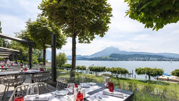 An outdoor dining area with tables set for two, overlooking a lake and mountains. - HERMITAGE Lake Lucerne - Beach Club & Lifestyle Hotel (Lucerne)