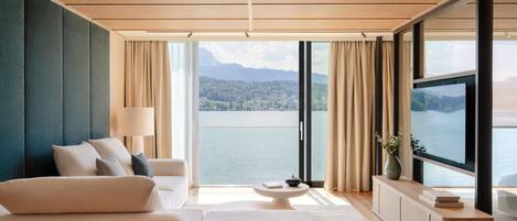 A modern hotel room with a large window offering a lake view, a sofa, a coffee table, and a TV on a cabinet.