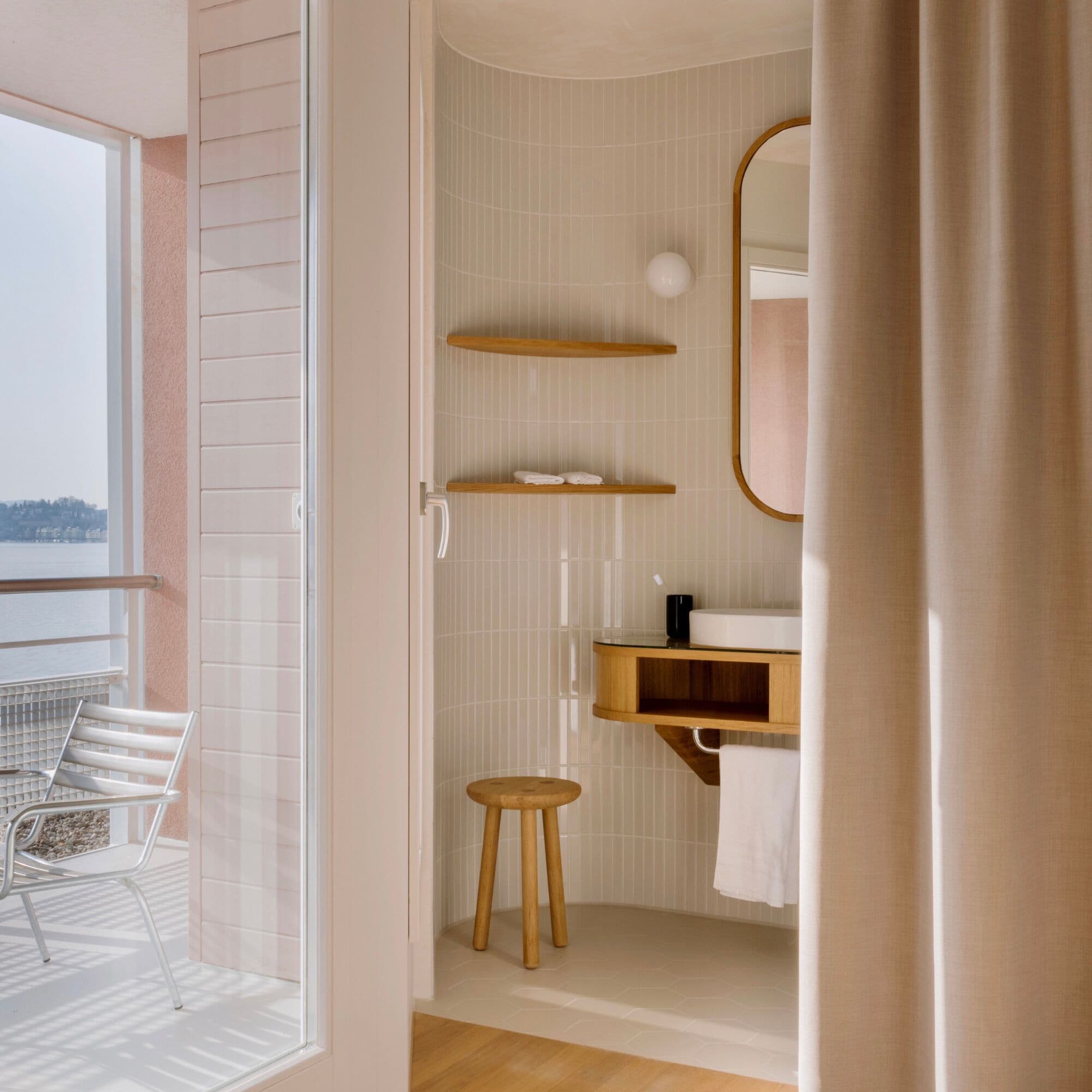 A bathroom with a shower, a toilet, a sink with a mirror, and a chair by a window.