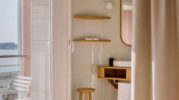 A bathroom with a shower, a toilet, a sink with a mirror, and a chair by a window.