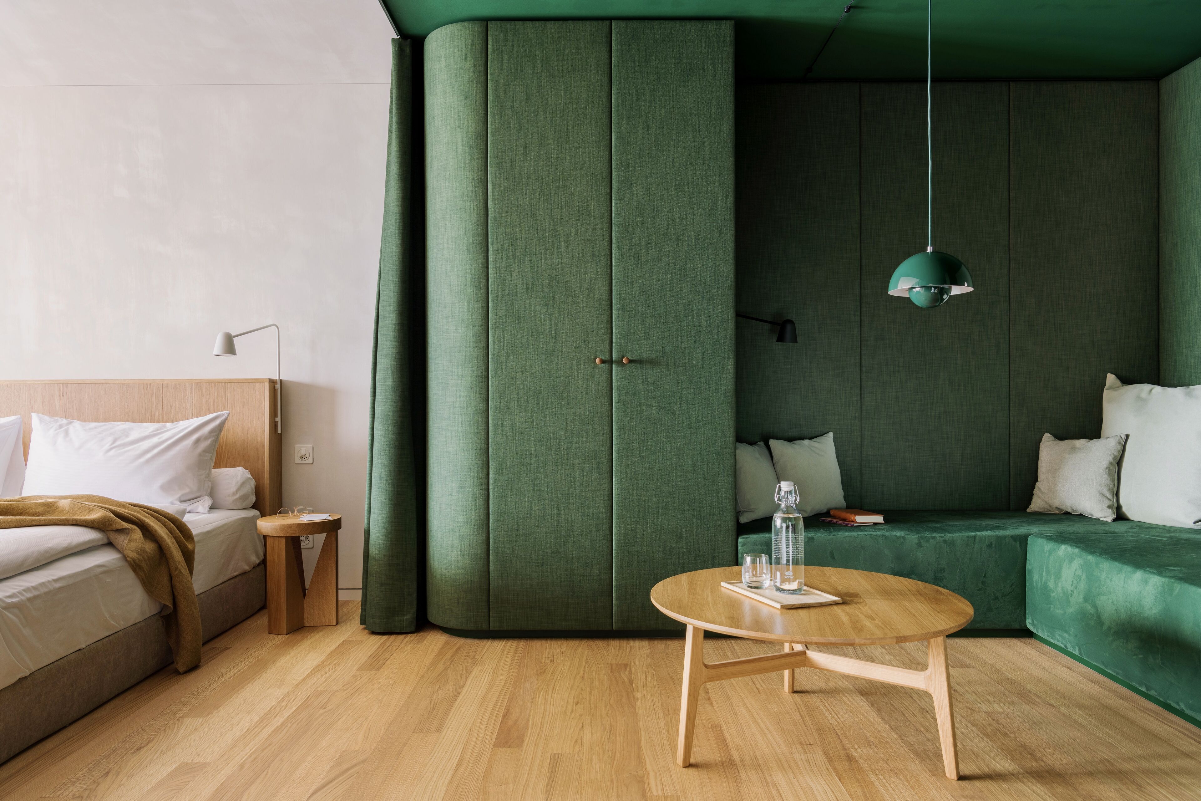 A modern hotel room with a bed, a small round table, a green sofa, and a green pendant light.