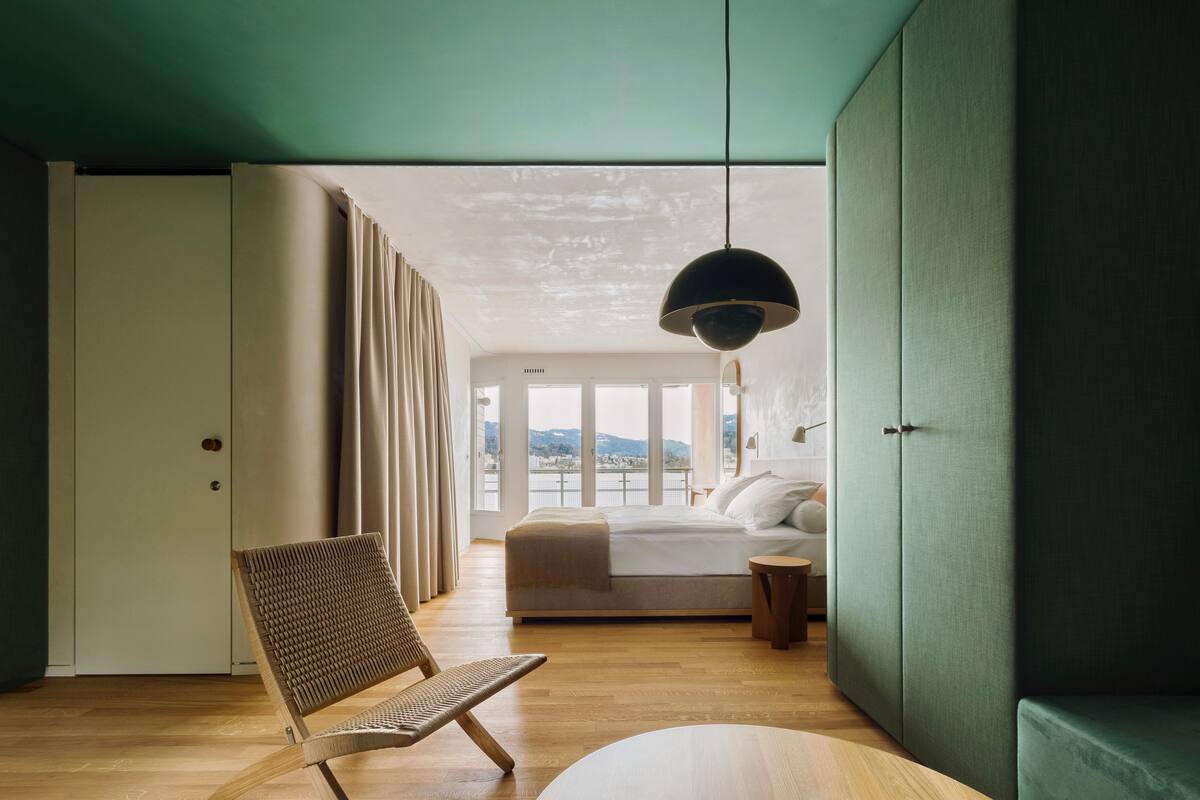 A bedroom with a bed, a chair, a bedside table, and a view of the outdoors.