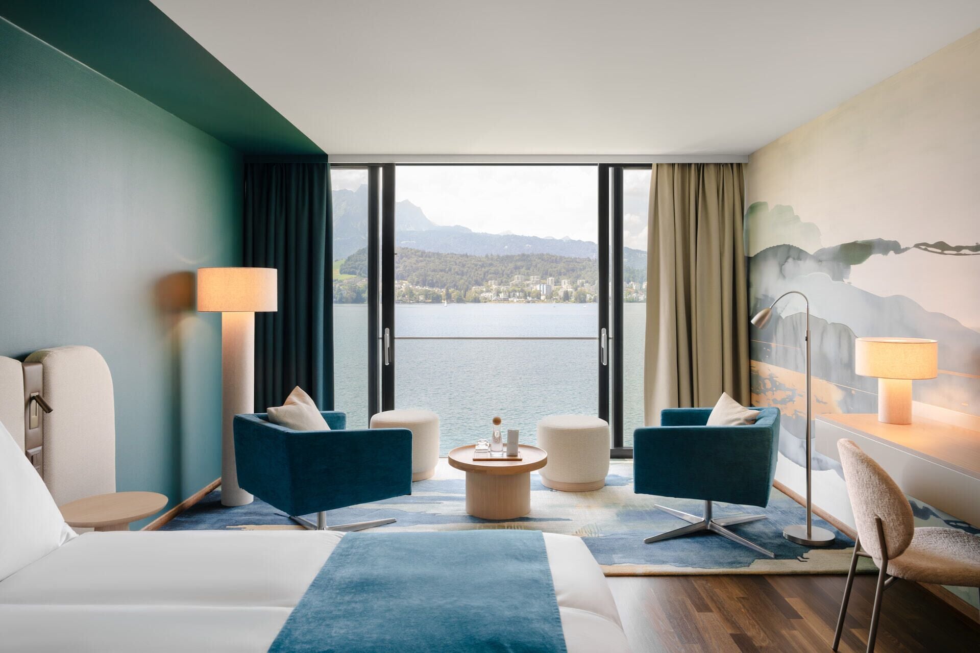 A hotel room with a large bed, two armchairs, a small round table, and a view of a lake and mountains.