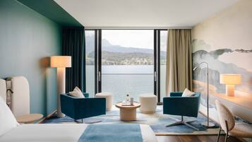 Habitación de hotel con una cama grande, dos sillones, una mesita redonda y vistas a un lago y montañas.