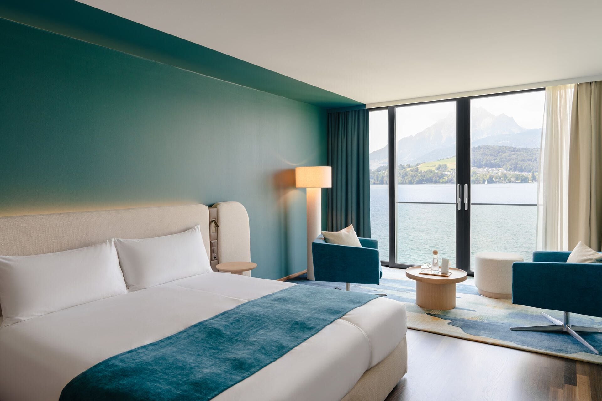 A hotel room with a large bed, a desk, two chairs, a lamp, and a view of a lake and mountains.