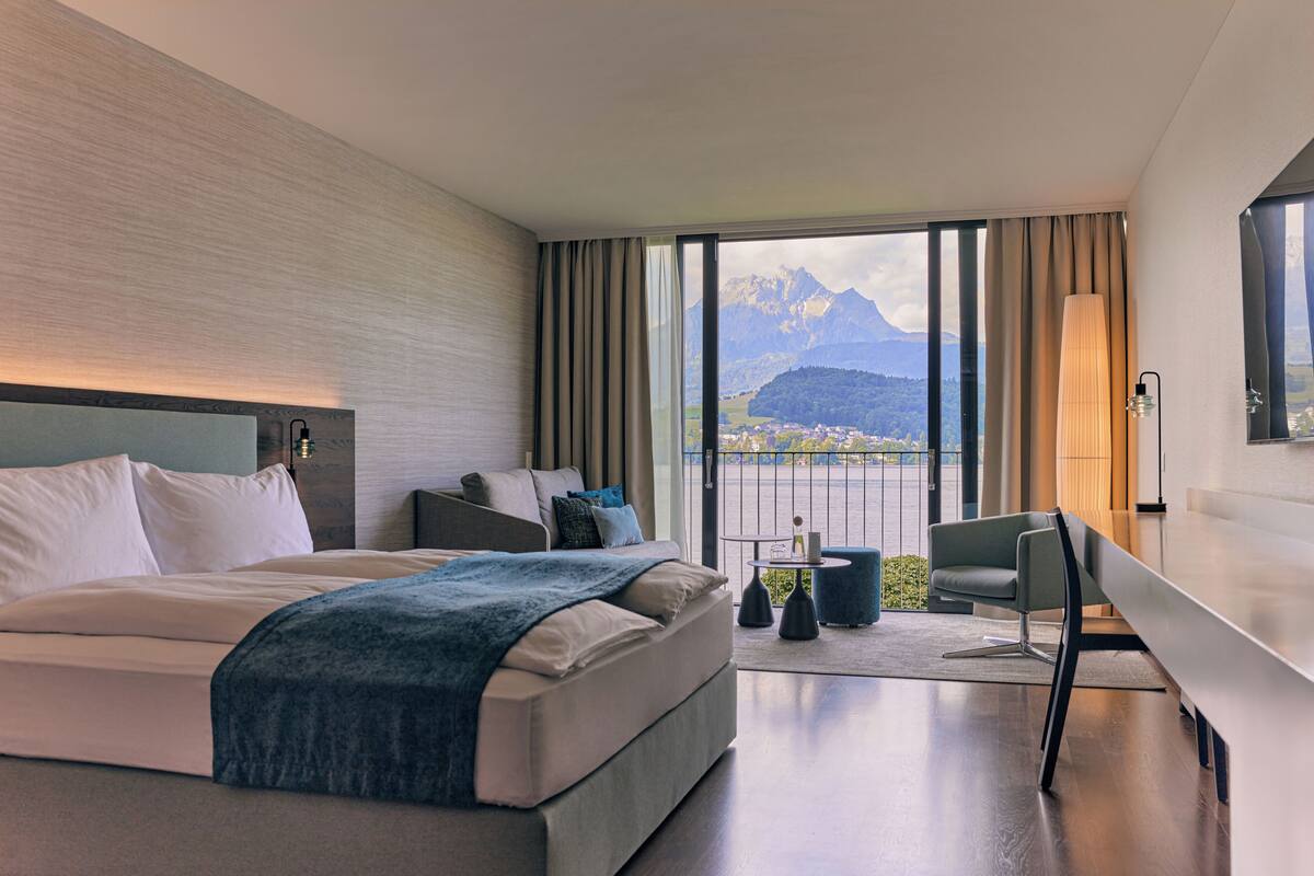 A modern hotel room with a large bed, a desk, a chair, and a view of mountains.