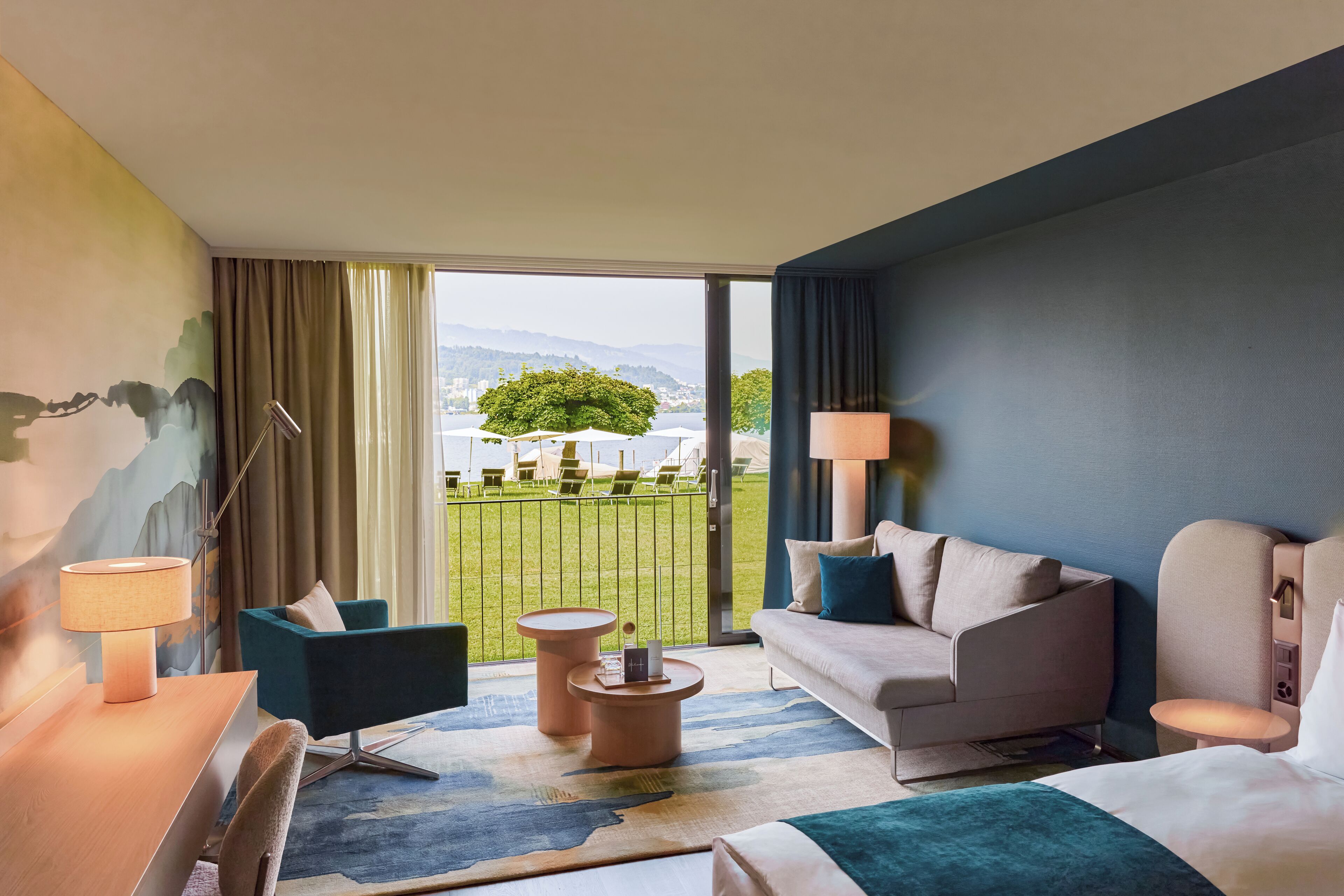 Una habitación de hotel con un ventanal grande, un sofá, una silla, una mesita y una lámpara.