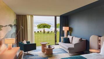 Una habitación de hotel con un ventanal grande, un sofá, una silla, una mesita y una lámpara.