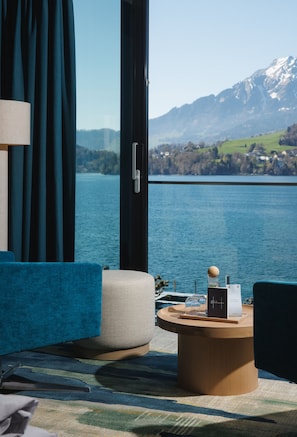 A room with a view of a lake and mountains, a blue sofa, a round wooden table with a glass, and a lamp.