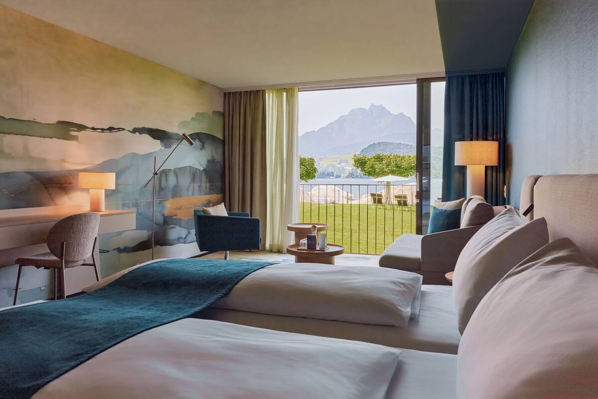 A hotel room with a large bed, a desk, a chair, a small table, and a view of mountains through the window.