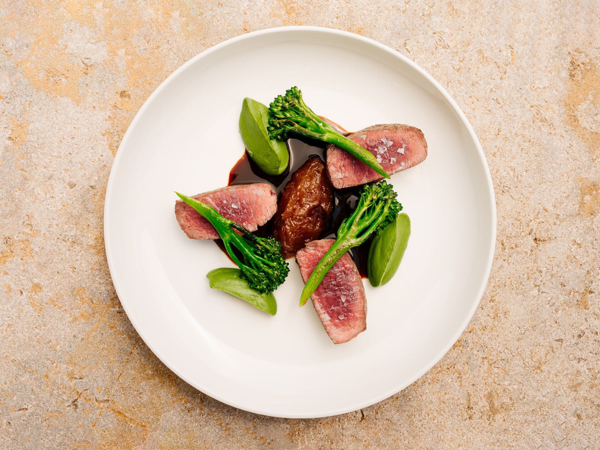 Un plato blanco con carne en rodajas, brócoli y un vegetal verde sobre una superficie rugosa.